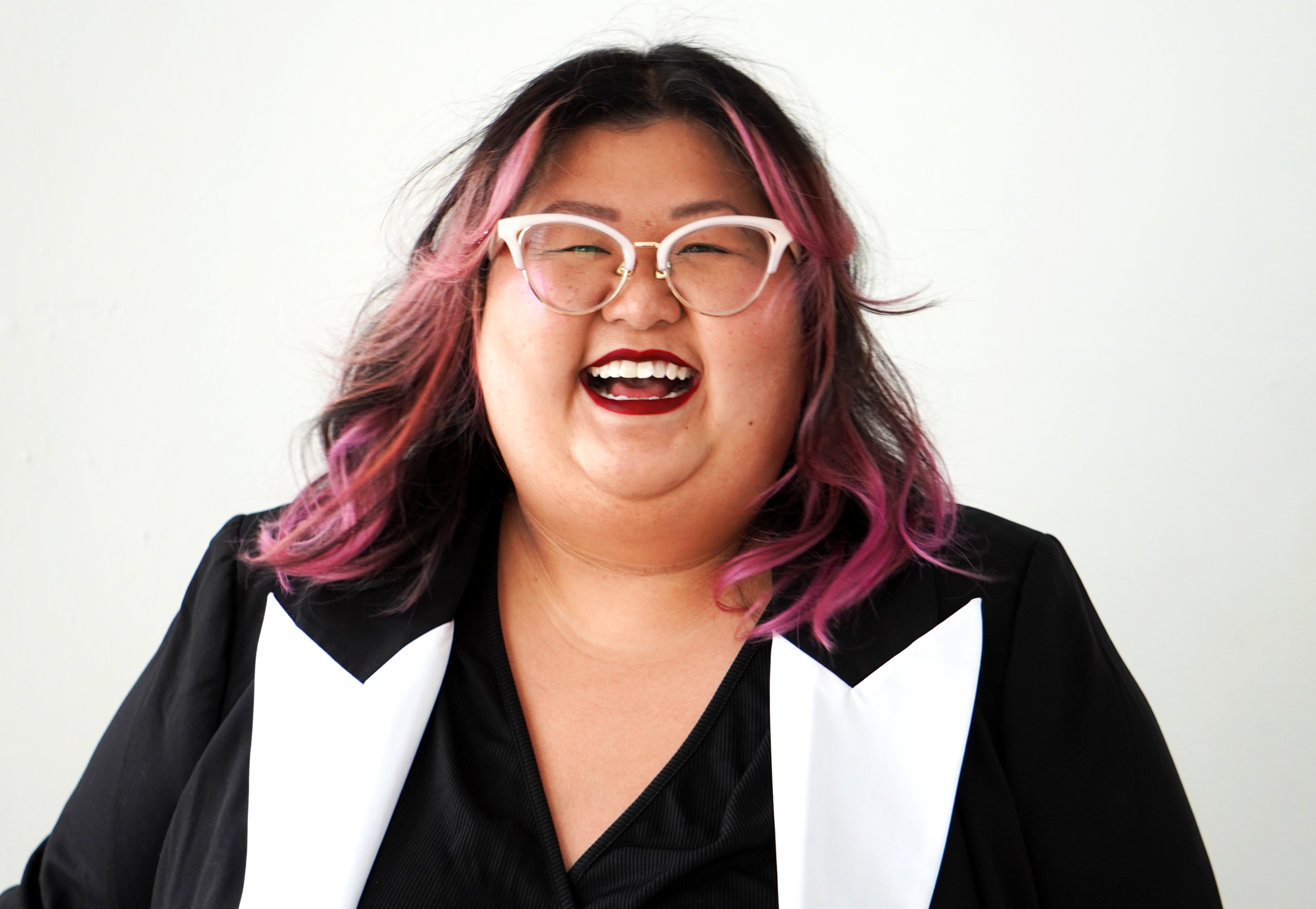 A smiling person of medium light skin tone with pink and black hair, wearing red lipstick and white rimmed cat eye glasses, and a black and white jacket.