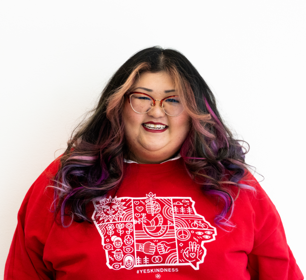 A smiling person of medium light skin tone, with pink, purple and black curled hair, wearing red lipstick and red rimmed cat eye glasses, and a red sweater with the outline of Iowa and various images drawn within it and the phrase: "#yeskindness"