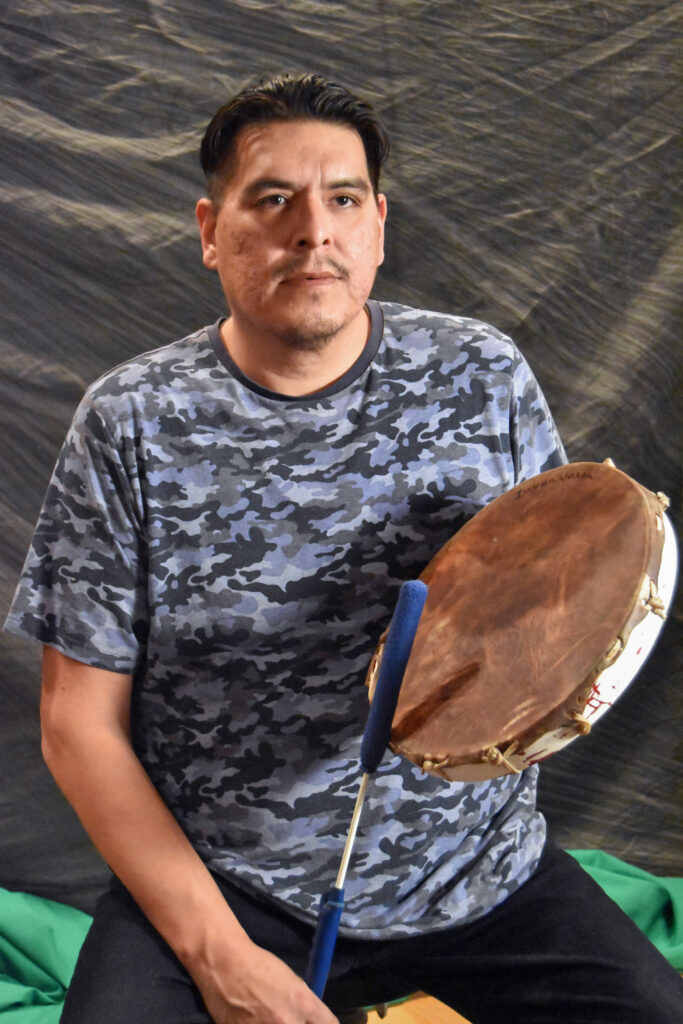 A man sitting, holding a traditional Native American percussion instrument with his hands.
