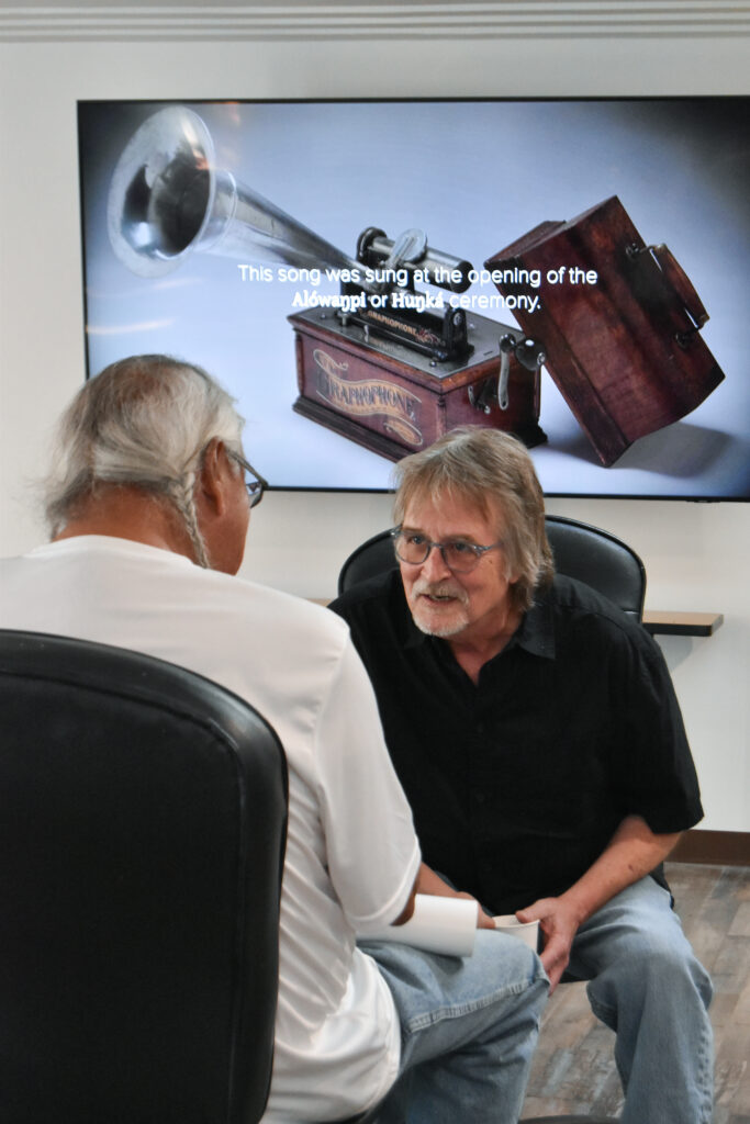 Two men sitting and talking. One in a white shirt has his back to the camera, while the other in a black shirt and glasses leans over to talk.