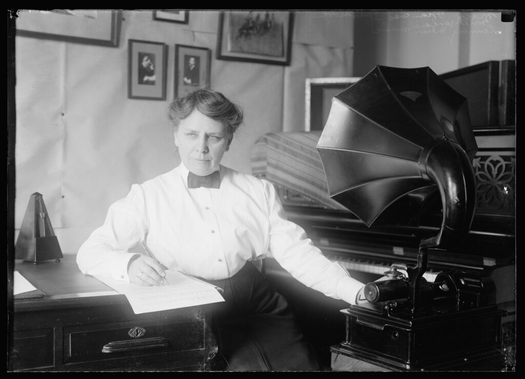 A woman sitting between two tables in the early 1900s. She's using one hand to write on a sheet of paper, with the other hand placed on a gramophone.