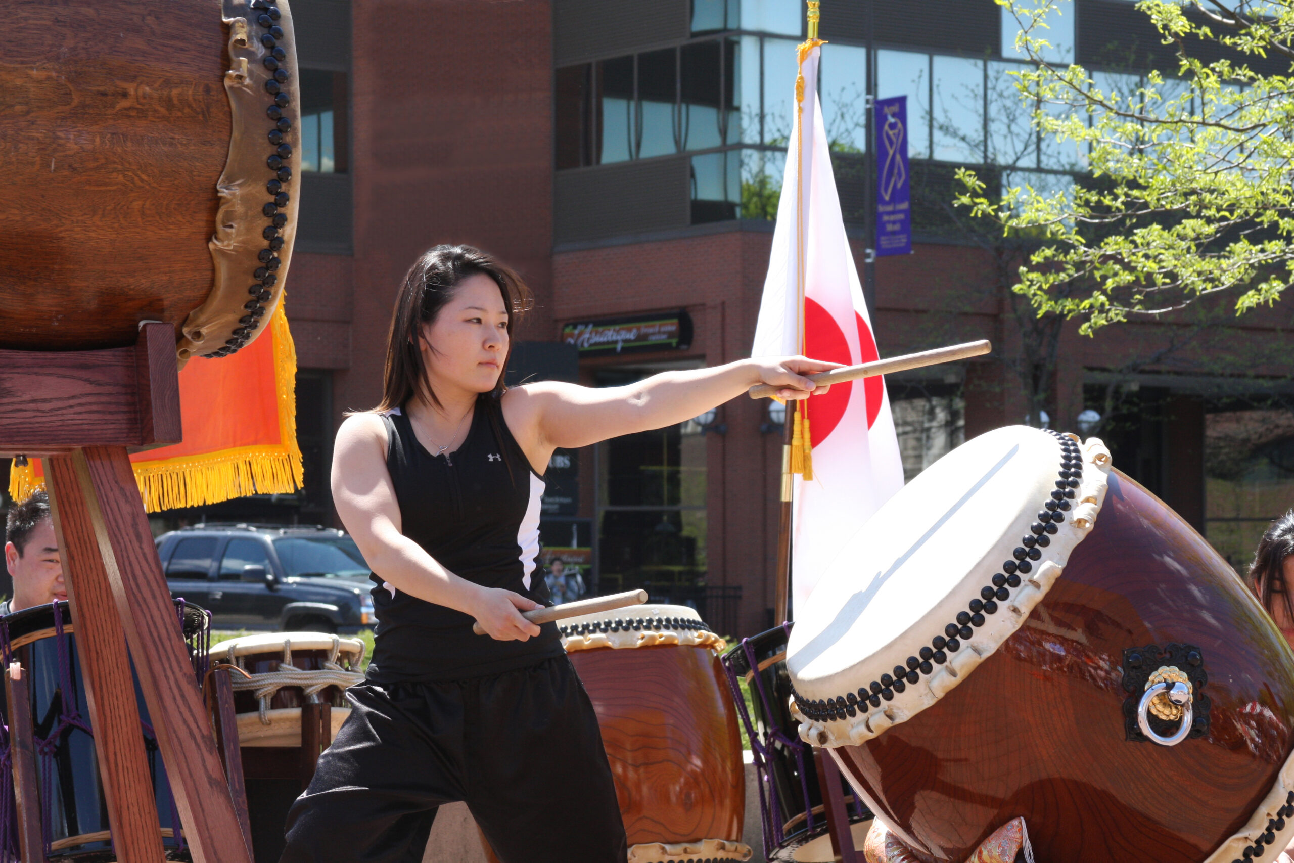 A person stands outside a building, surrounded by various drums, posing with arms out to one side and holding drumsticks.