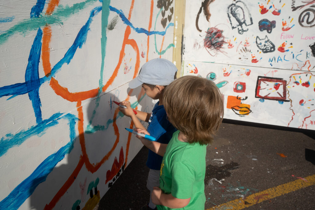 Two young children draw with markers on a standing drywall filled with other illustrations as part of an outdoor community art event at Innoskate 2022 in downtown Sioux Falls, SD.