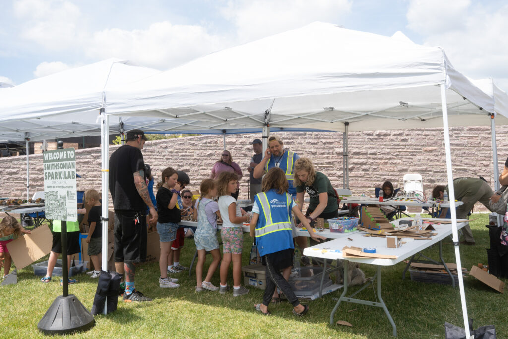 A group of young children gather under a white tent to be assisted by adults to participate in an activity during Innoskate 2022 in downtown Sioux Falls, SD. There is a sign beside the outdoor tent that reads, 'Welcome to Smithsonian Spark! Lab.'