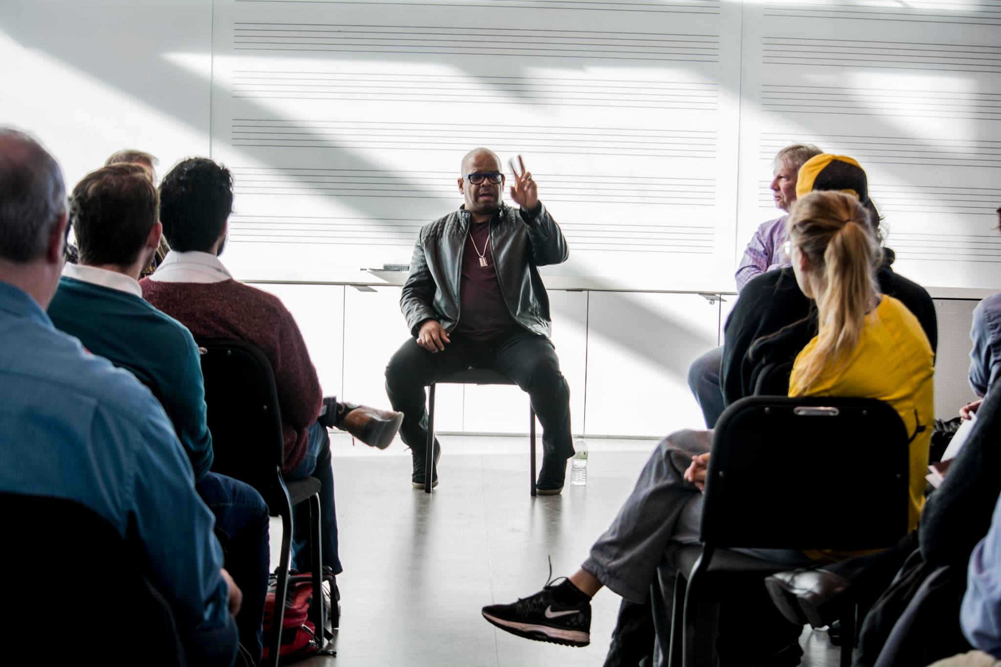 A person sits at the front of a room before a seated audience, holding two fingers in the air and talking.