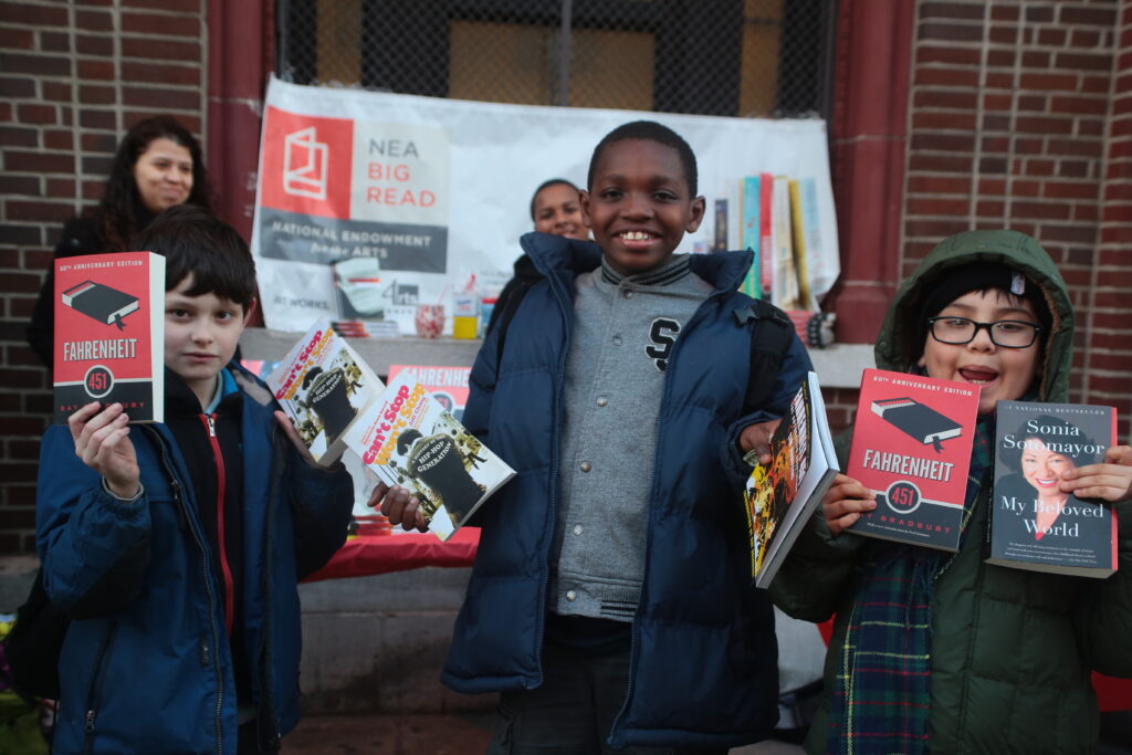 Three young people pose with books, including "Fahrenheit 451," "My Beloved World," and "Can't Stop Won't Stop," in front of an NEA Big Read banner.