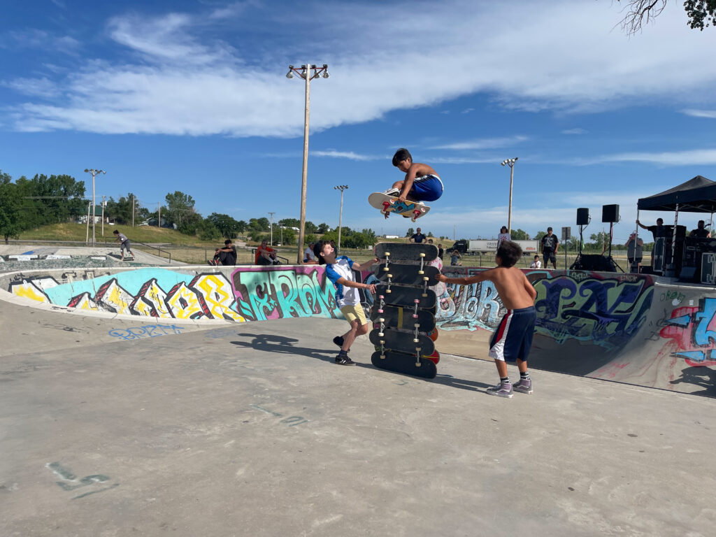 Two young skateboarders support a stacked tower of seven skateboards while another young skateboarder is mid-air, jumping over them on his board, at a cement skatepark in Pine Ridge Reservation, South Dakota.