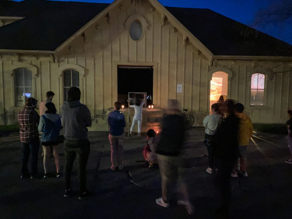 About a dozen people stand around and watch a performance of a person wearing white outside a yellow cottage-like building with soft light shining through the windows into the outdoor evening event.