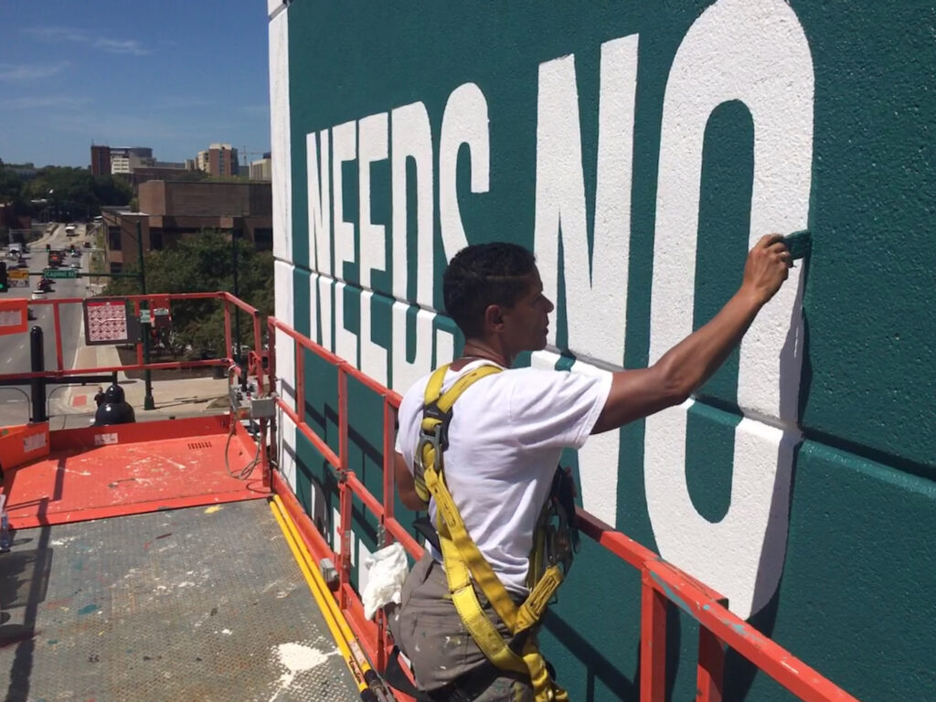 A person wearing a yellow harness on a big lift stands and paints the side of a building with letters that spell "Needs No...".