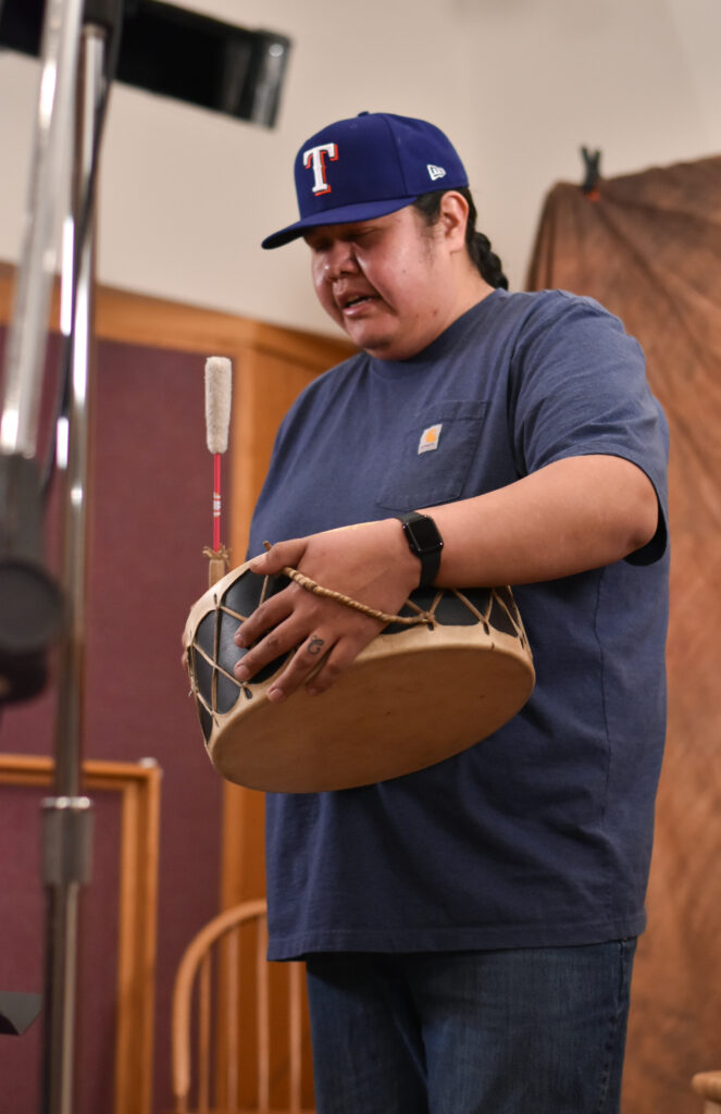 A man in a blue shirt and baseball shirt sings and plays a traditional Native American handheld percussion instrument.
