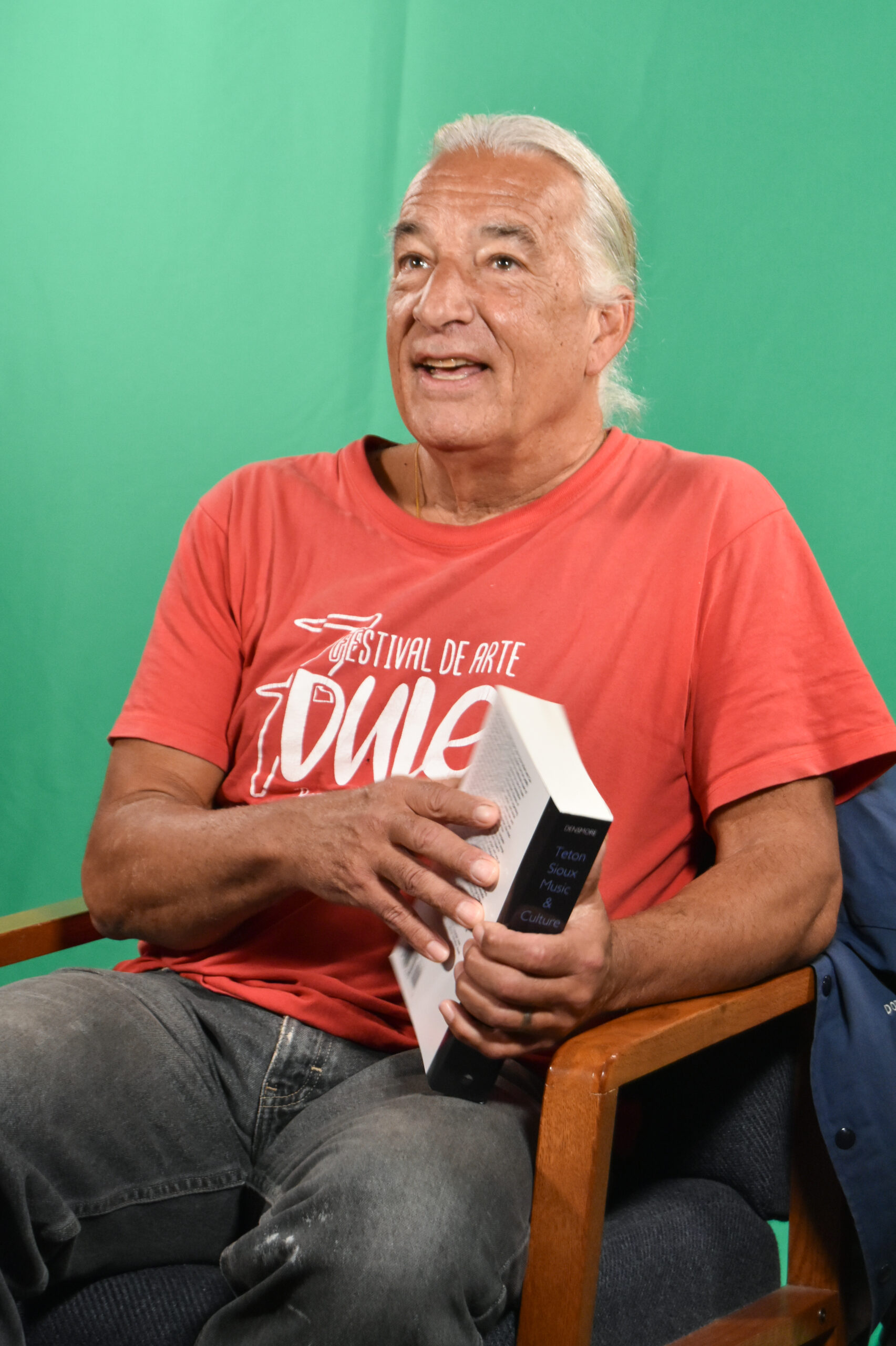A Lakota man with white hair, wearing a red T-shirt and black jeans sitting on a wooden armchair. He is holding a thick book while captured in this photo mid-sentence.