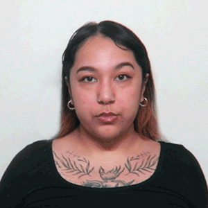 Gif of two interchanging headshots of a person of medium light skin tone, long dark hair, wearing small hoop earrings and a black shirt with a tattoo above the neckline. In one headshot their hair is down, in the other it's pulled back.