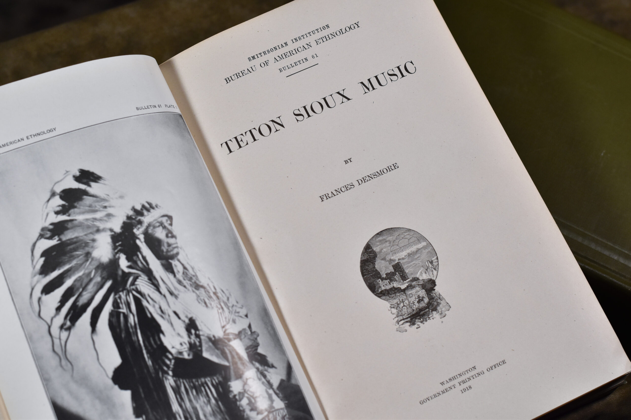 An open book with apage showing the title, Teton Sioux Music by Frances Densmore, and a facing page containing a photo of a Native American person in a traditional headgear.