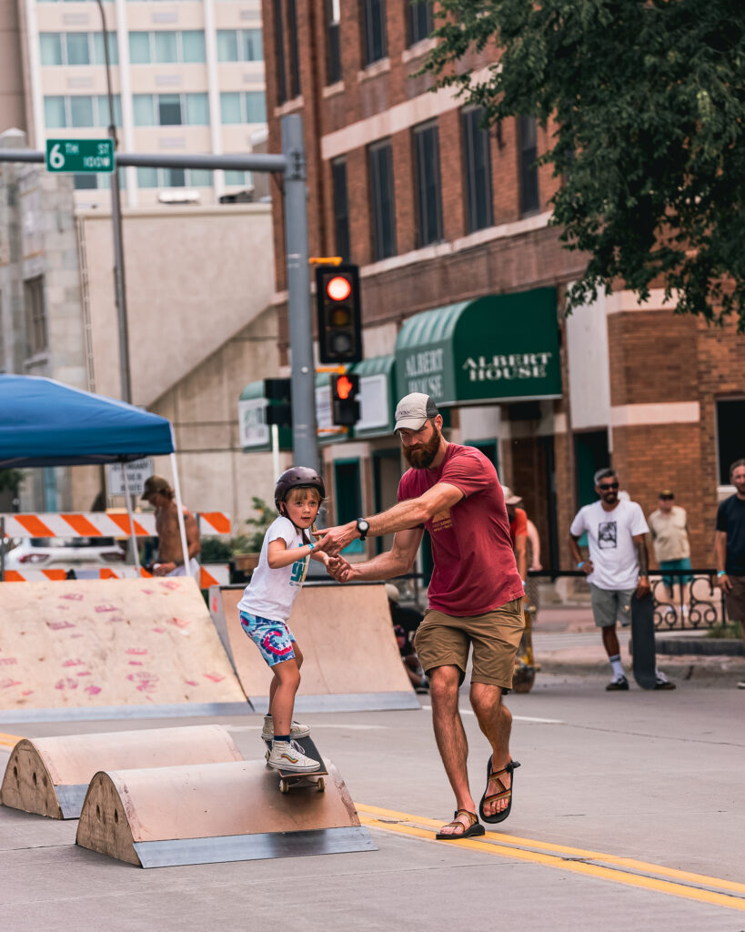 An adult helps a young skateboarder over a small ramp by holding their hands during Innoskate 2022 in downtown Sioux Falls, SD.