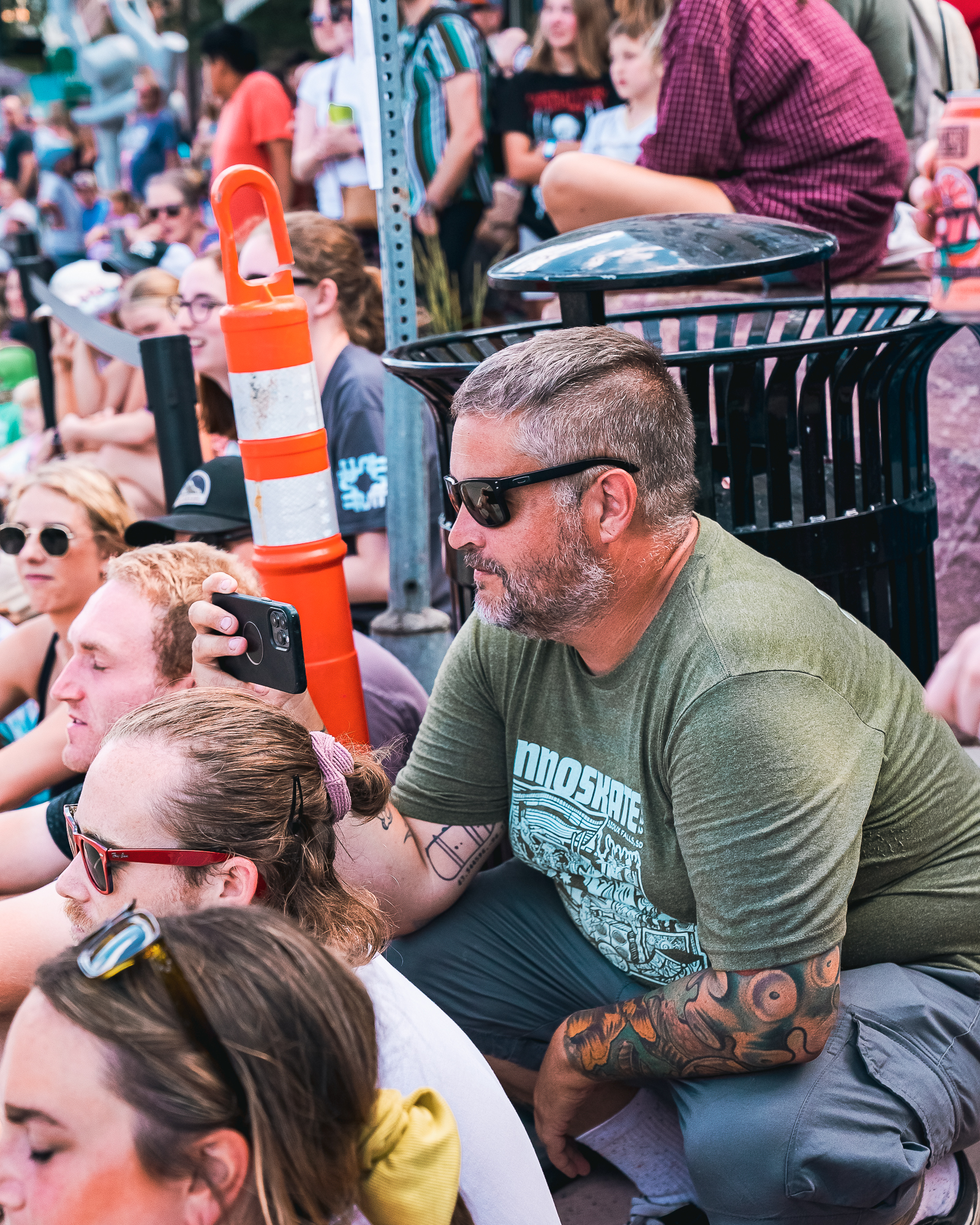 A person wearing black sunglasses squats holding a phone to film a video while surrounded by a crowd of people watching skate contests during Innoskate 2022 in downtown Sioux Falls, SD.