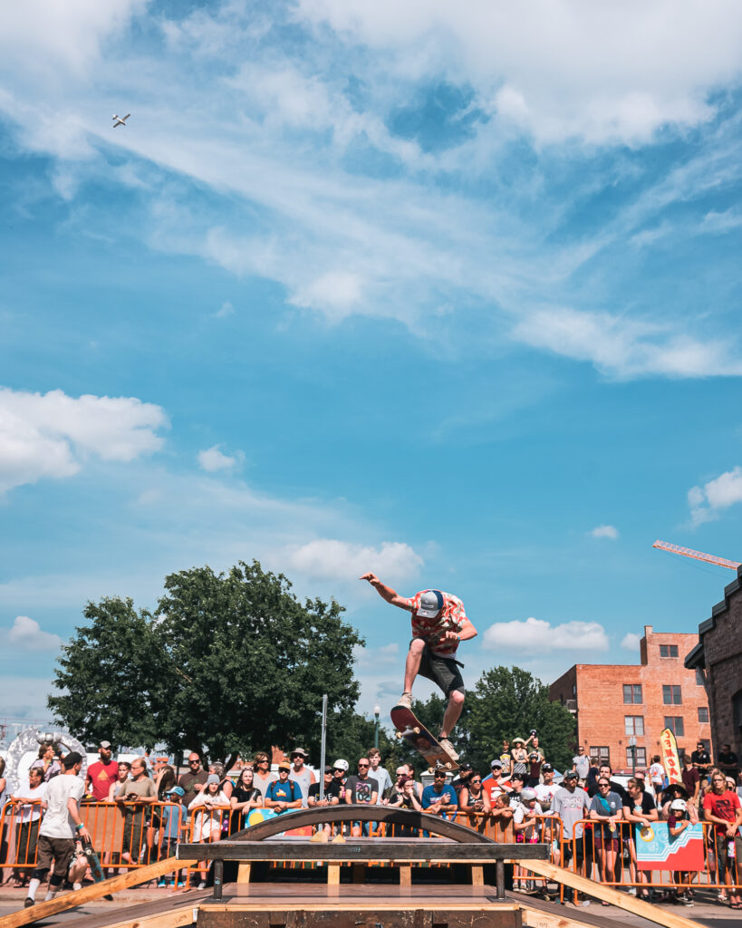 A skateboarder lifts off a skate ramp with his board and arms in the air during a sunny day with blue skies at an event called Innoskate 2022 in Sioux Falls, SD. There is a crowd of people standing in the background, behind a barricade watching the skateboarder.