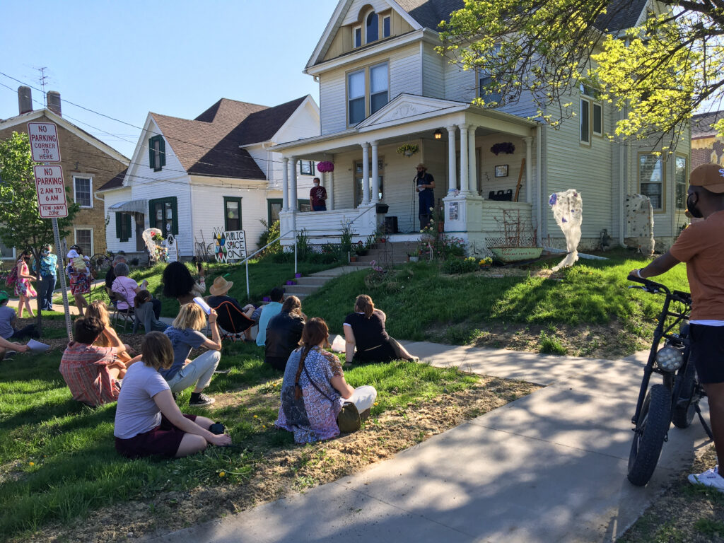 A seated audience on grassy ground watch a performer sitting in front of them on a porch of a white two-story building.