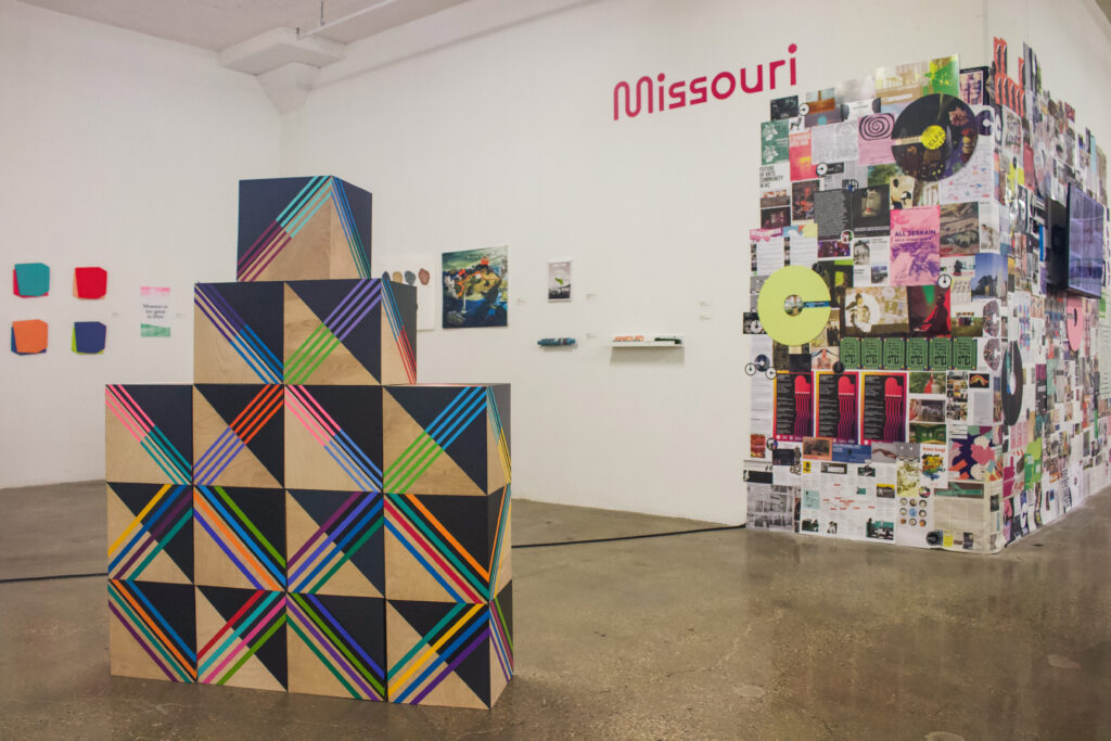 An installation of stacked cube boxes made of wood and painted with black colors and colorful lines. In the background, white gallery walls are covered with a colors of paper, two-dimensional artworks that wrap and extend around the corner of a wall.