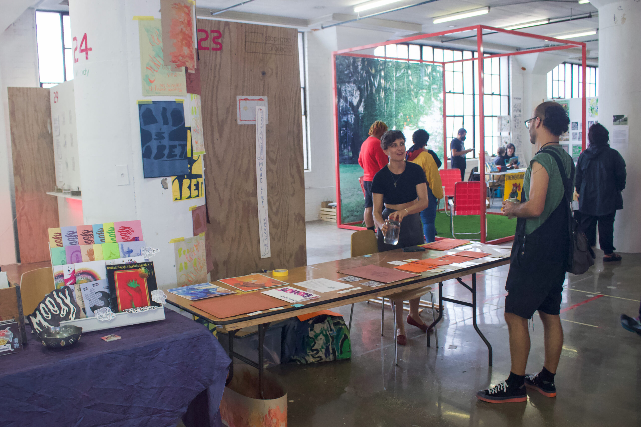 Two people standing and talking to each other across from a long table with printed art and publications for display. In the background, there is an art installation framed by a red cube with people standing around it.