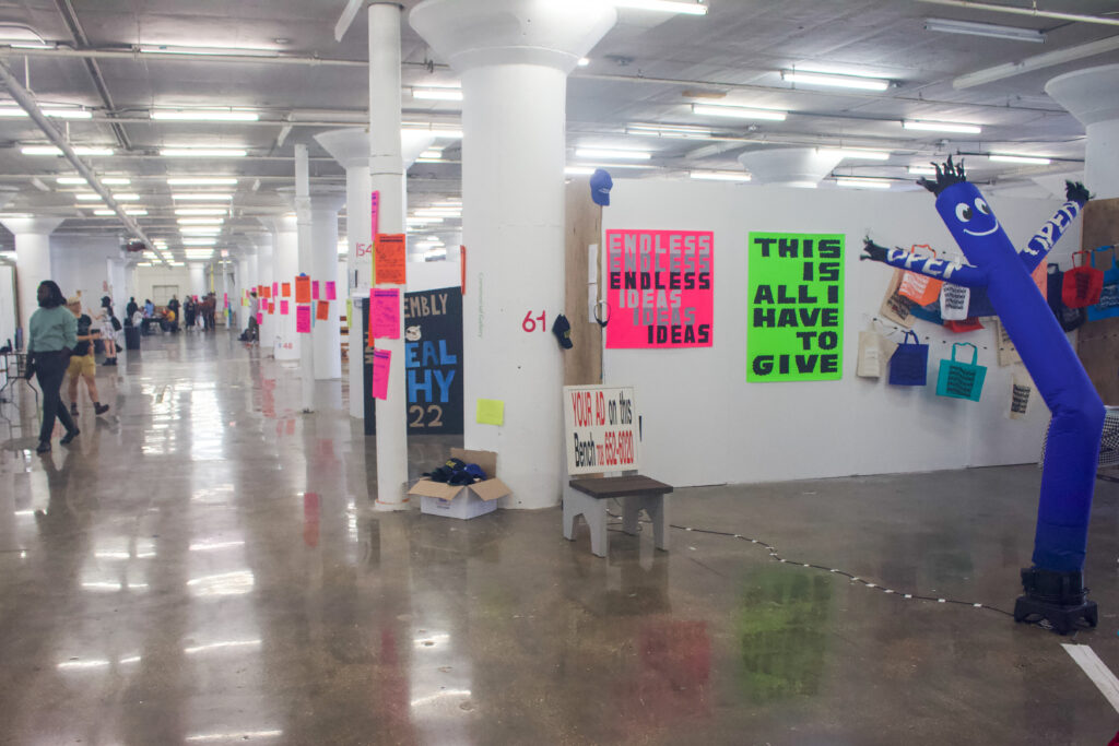 A wide shot of a floor showing a row of booths separated by white walls at the MdW Assembly held at Mana Contemporary Chicago. The floor has pillars covered in bright pink posters. At the edge of the frame, there is a bright blue inflatable figure with "open" written on its arms.