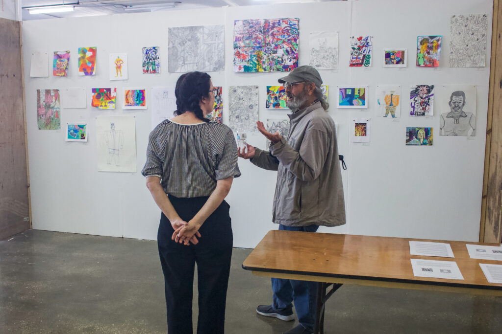 Two people talk while standing next to a table in front of a white wall covered in colorful works on paper of varying sizes. The person on the right of the frame is gesturing with both their arms lifted and palms facing up, while the other person has their back to the camera and their hands behind their back.
