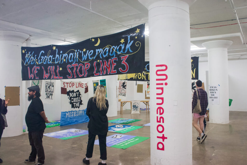 An area with white walls and a large pillar with pink lettering that reads, "Minnesota." There is a black banner hung across near the ceiling that with blue and red letters that reads, "Nibi Gaa-bimaji'we magak" and "we will stop line 3." The area and the floor have other protest posters.