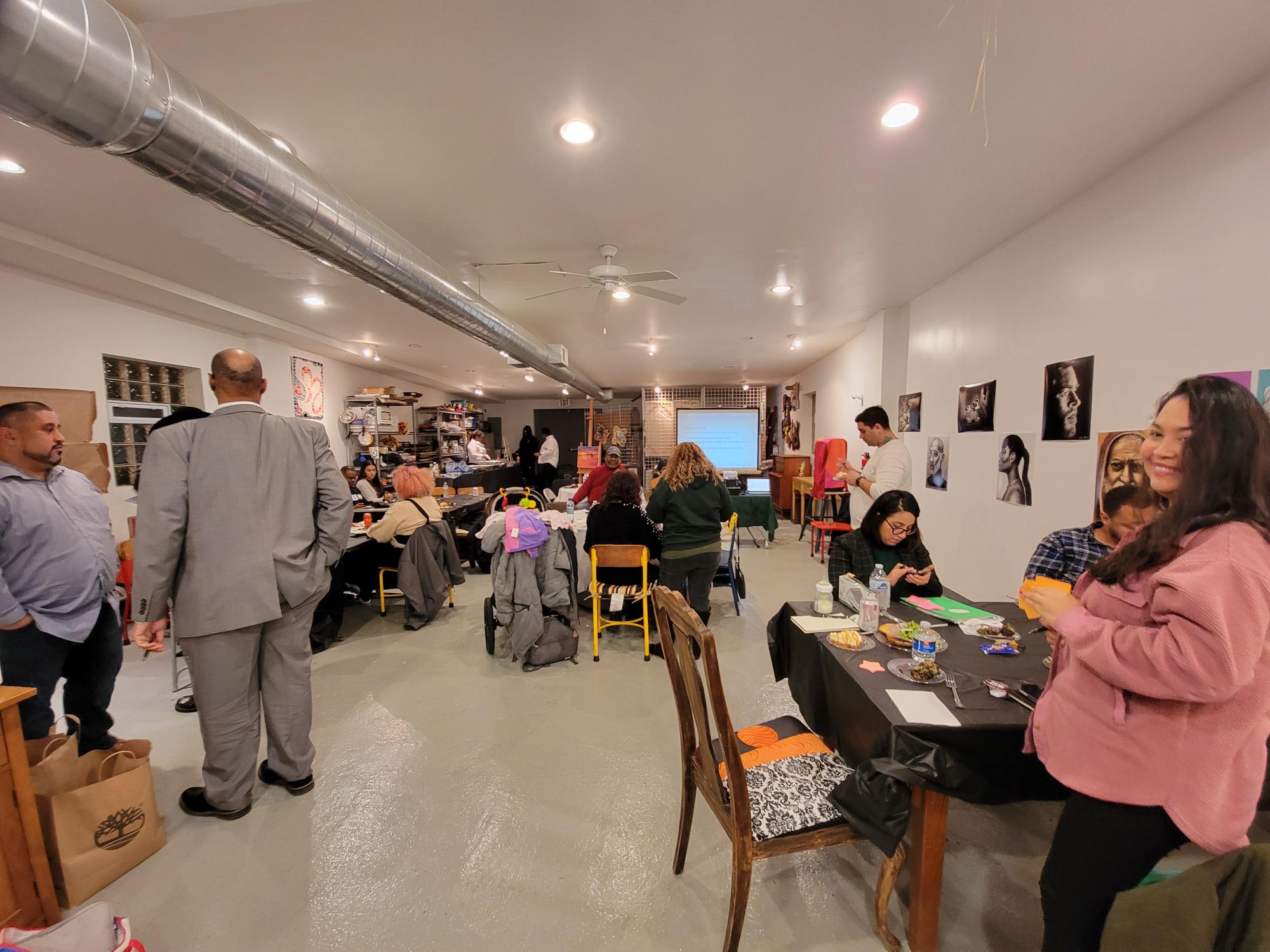 People of many ages and ethnicities gather in a multi-use art and craft space. A person close to the camera smiles while hoding a stack of sticky notes