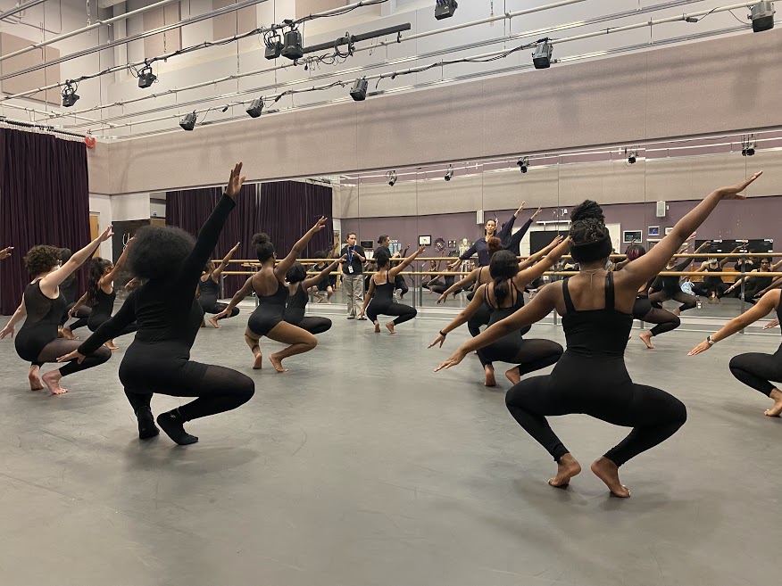 A group of Black dancers in a dance classroom crouch low to the ground with arms extended at an angle