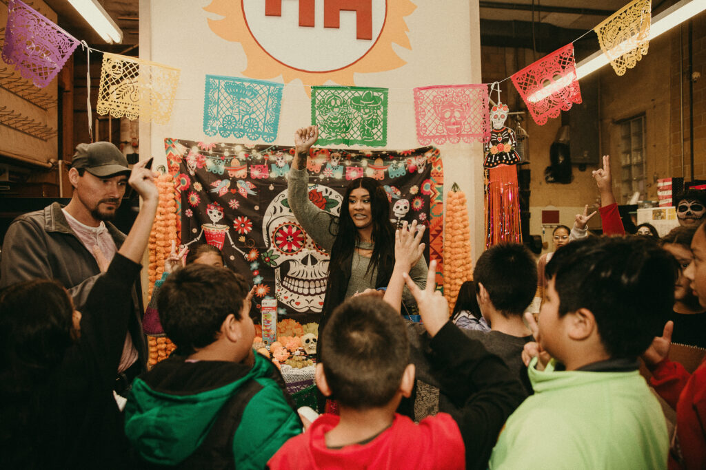 A group of young people hold up their hands while an adult holds up their fist at a at Dia de Los Muertos cultural celebration.