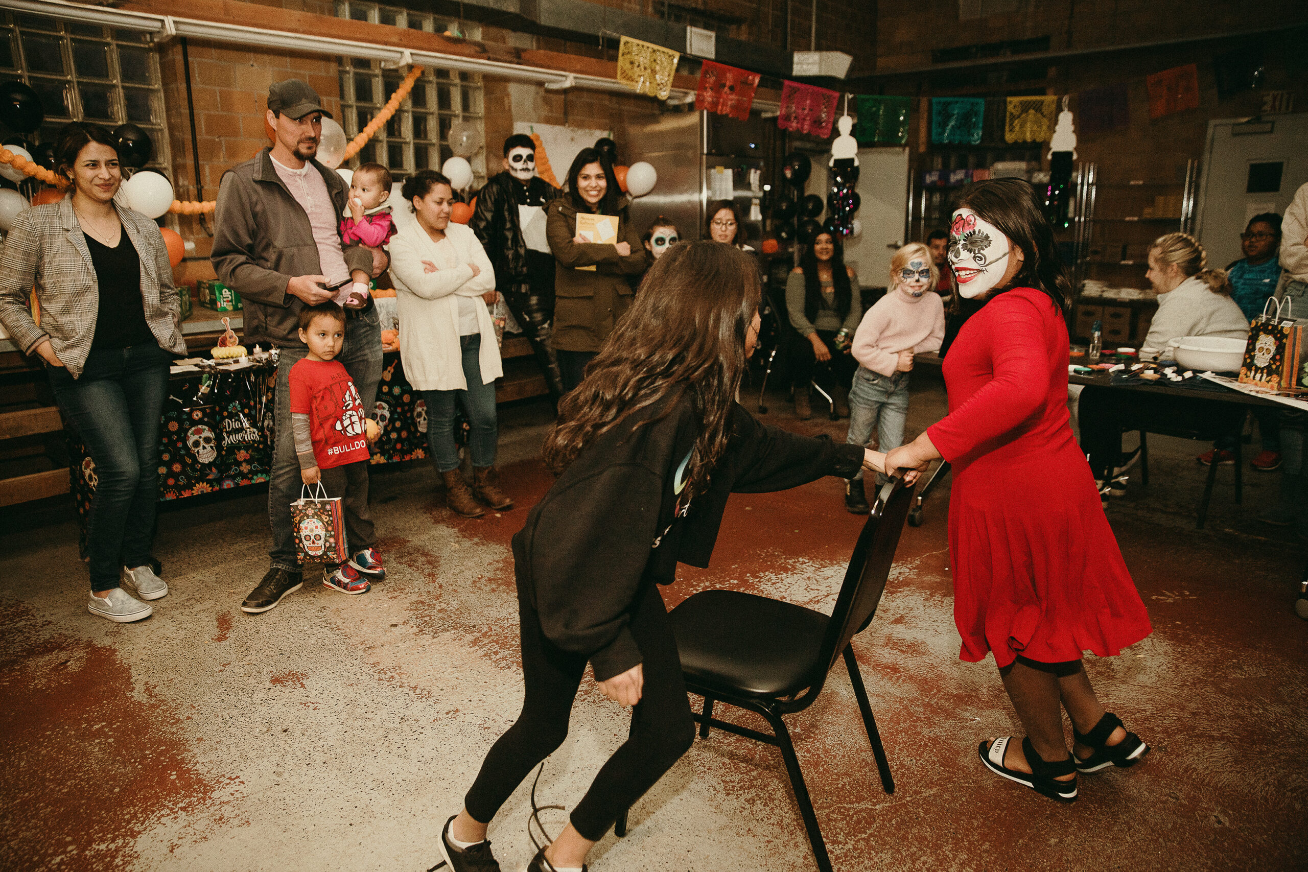 Two young people dance around a chair as others gather around them, as part of Dia de Los Muertos cultural celebration at La Luz Centro Cultural.