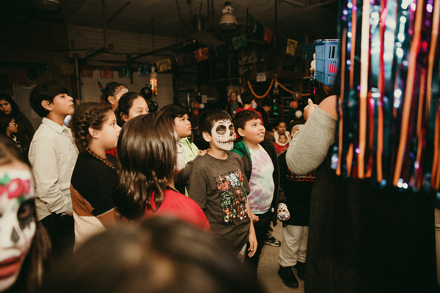 A group of young people stand in front of a offrenda at a at Dia de Los Muertos cultural celebration. A yound boy in the front has his head painted as a skill.