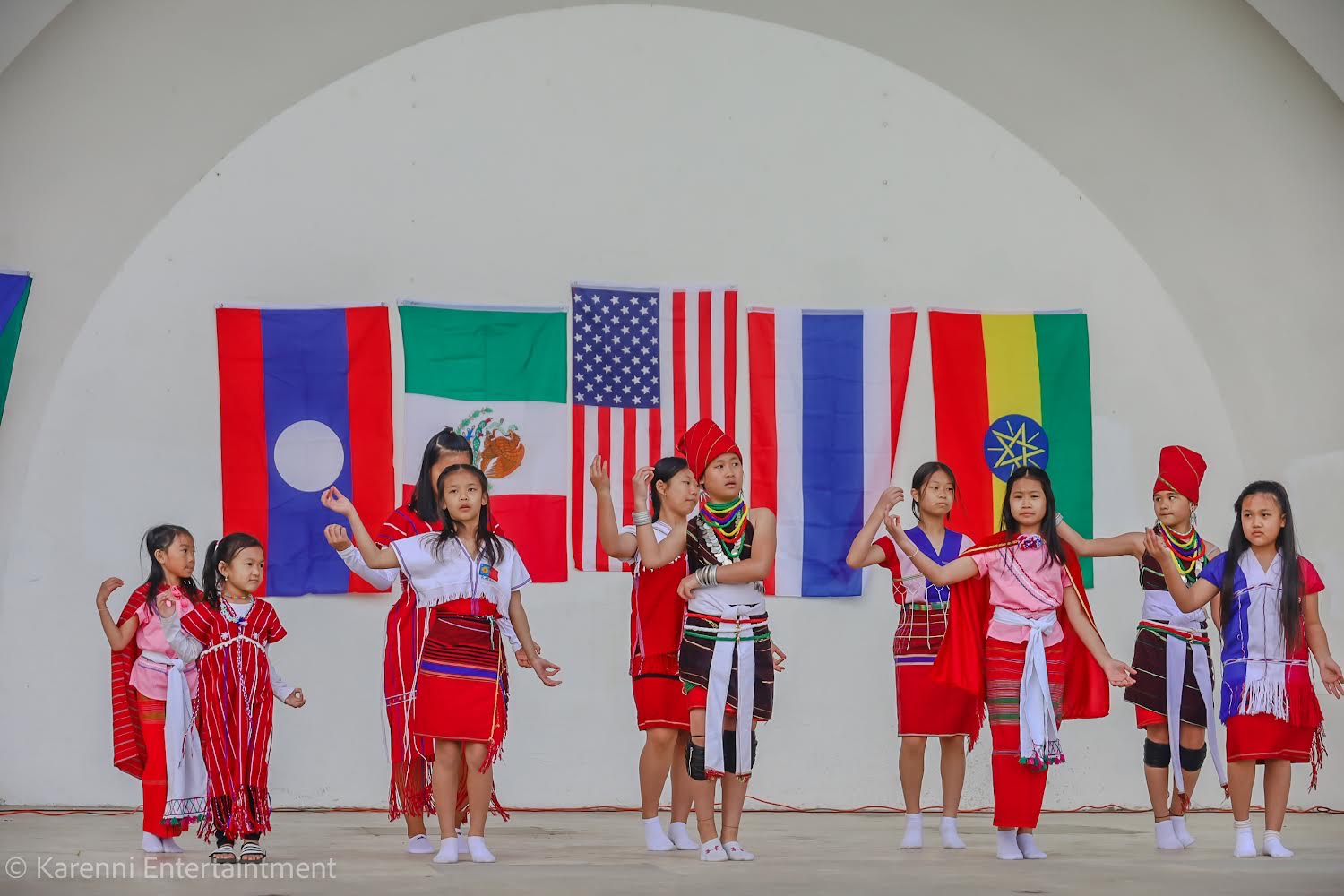 Ten children in traditional Hmong clothing dancing in front of a wall with flags of various countries.