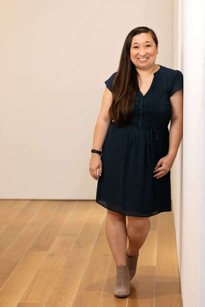 A full body headshot of a smiling person of medium light skin tone, with long dark brown hair tossed to the side, wearing a black dress and light brown boots, standing on hardwood floor in front of an off-white wall