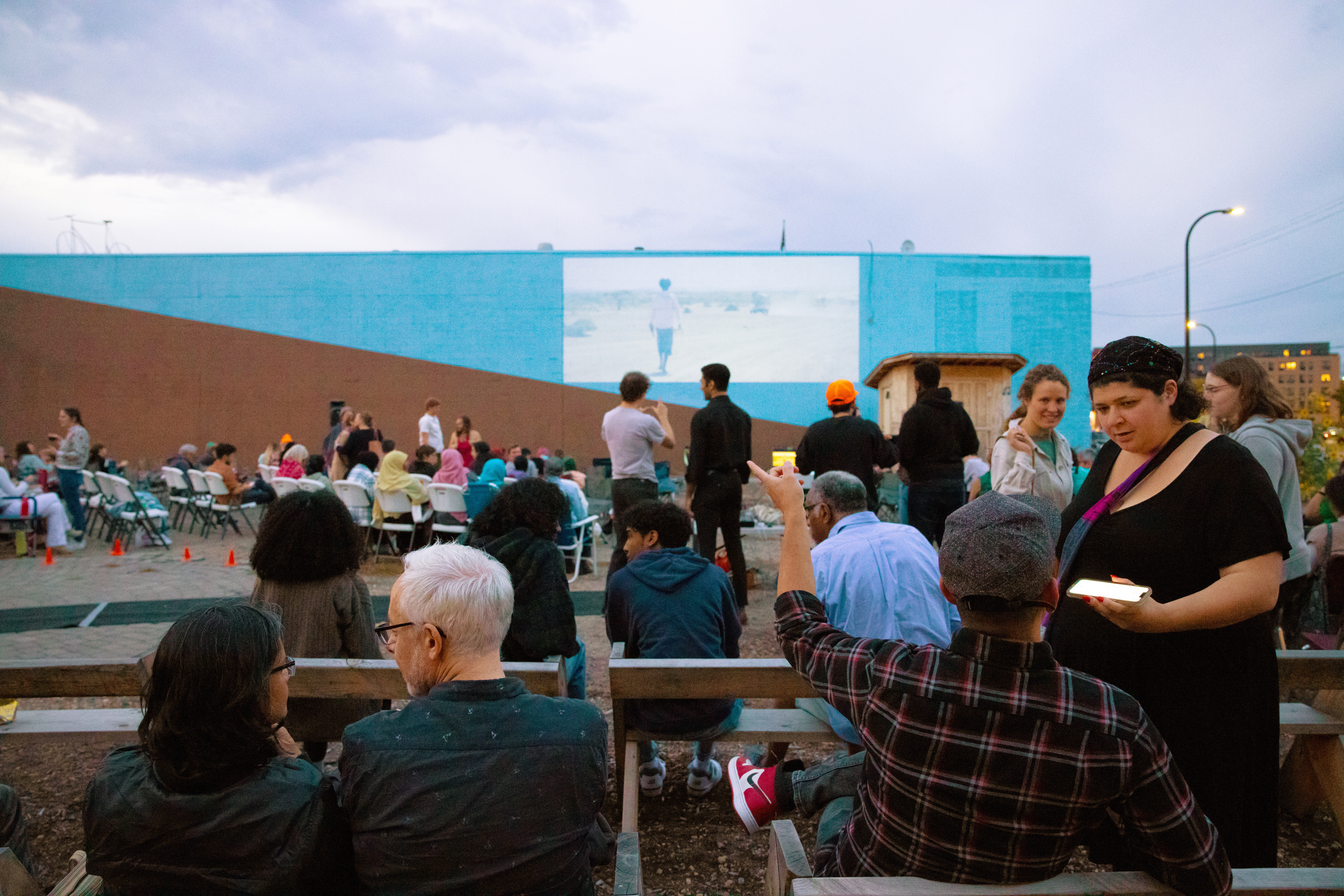 People sit around on wooden benches and in lawn chairs facing a tall side of a building that has a projected image during the 16th Arab Film Fest organized by Mizna in 2022 in Minneapolis, Minnesota.