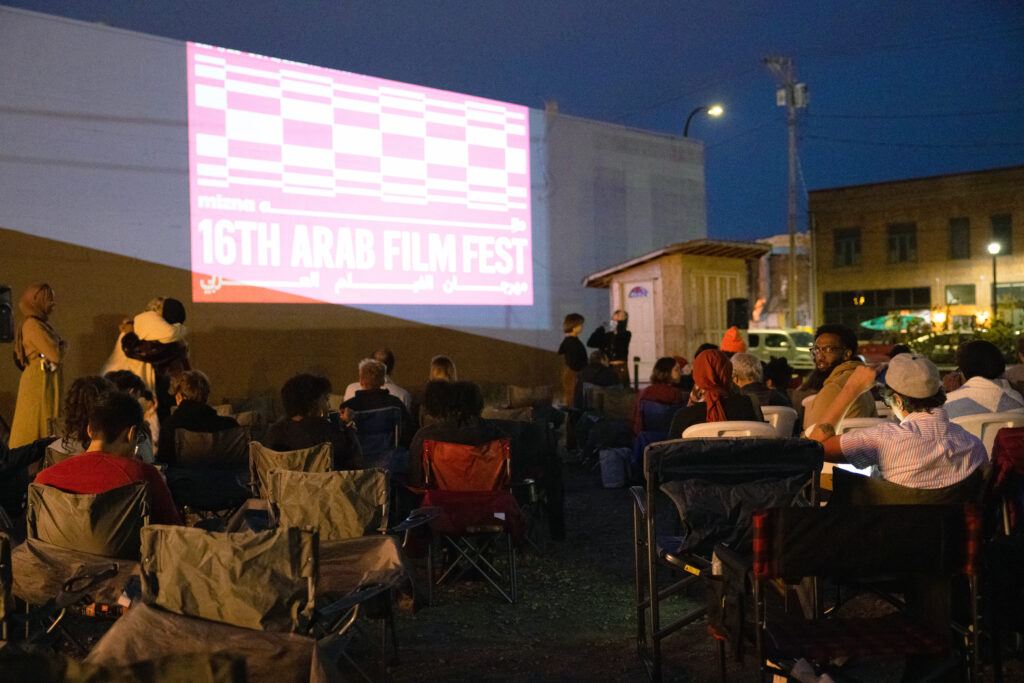 People sit around in lawn chairs facing a tall side of a building that has a projected image that reads "16th Arab Film Fest" as part of Mizna's 2022 event in Minneapolis, Minnesota.