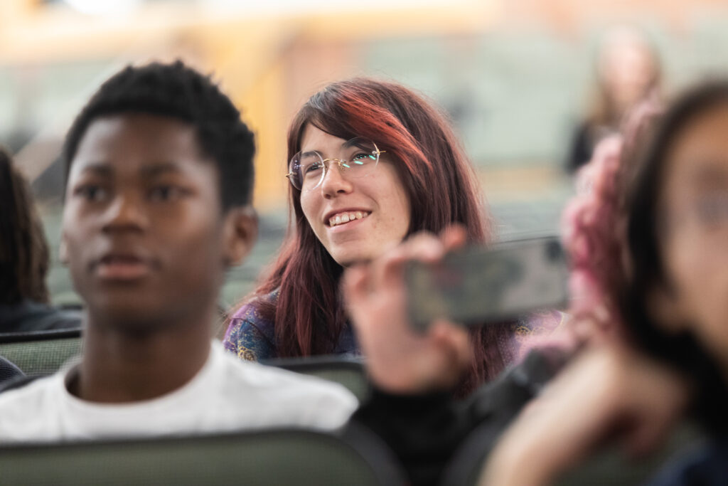 A smiling person with circular glasses and pink and brown hair sits between others in auditorium seats