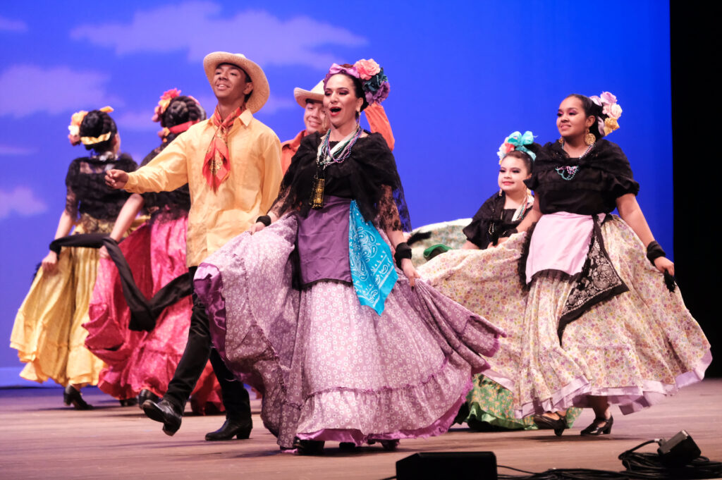 A group of dancers in colorful Baile folklórico costumes strut across a stage.