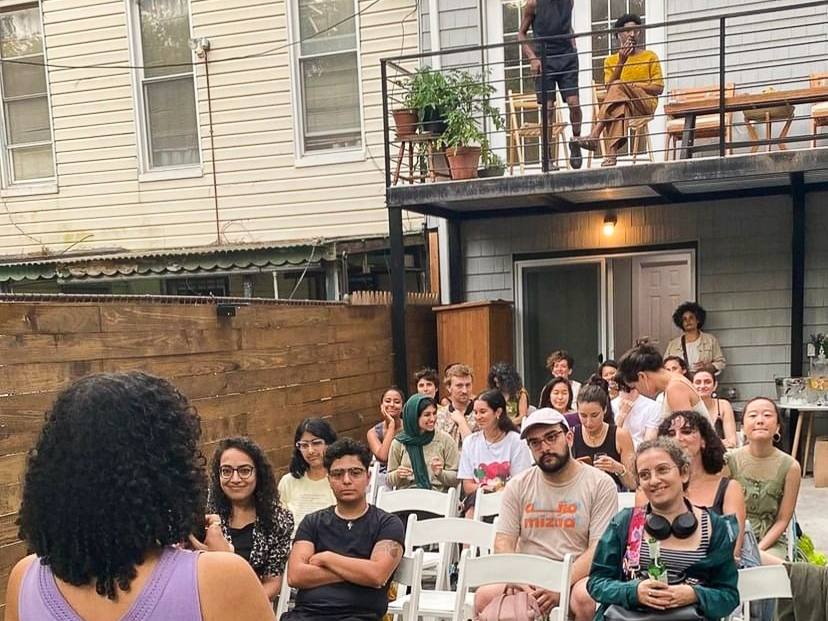 A seated audience in the backyard of a building listen to a literary reading.