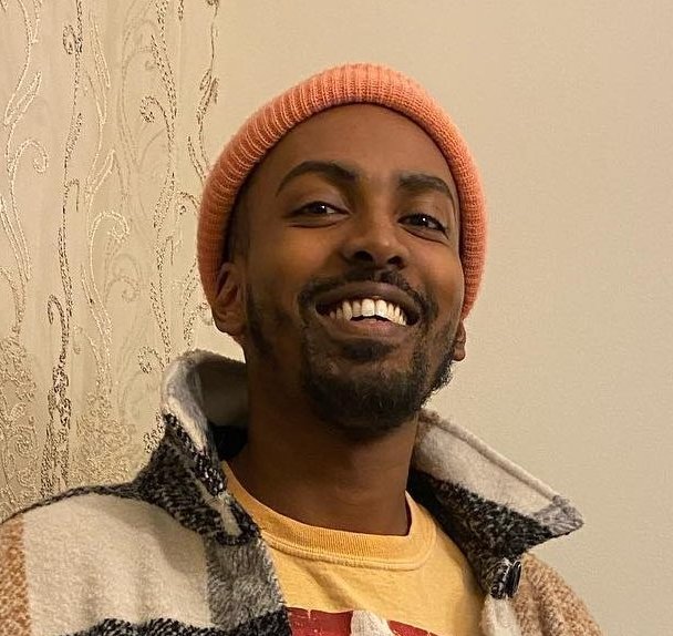 A smiling person of dark skin tone with trim black facial hair, wearing a salmon pink beanie and a flannel jacket, stands in front of a white wall