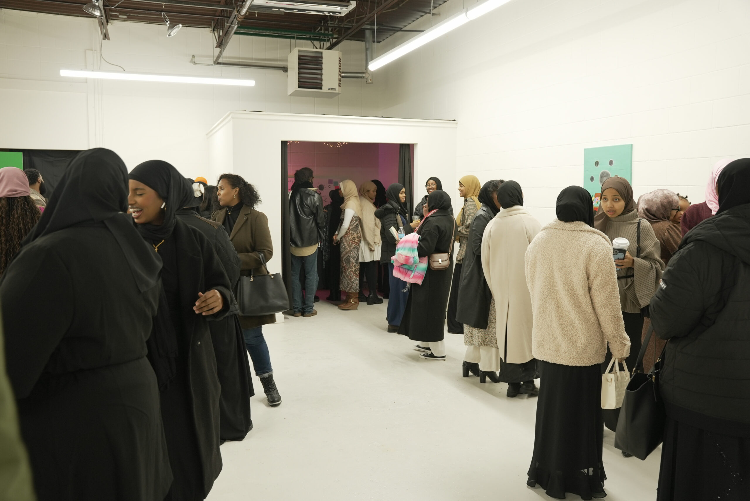 Attendees gather at the center of the gallery space following an artist talk at Soomaal House in Minneapolis, MN. The white walls, in frame, surrounding them have small to medium-sized two-dimensional works of art.