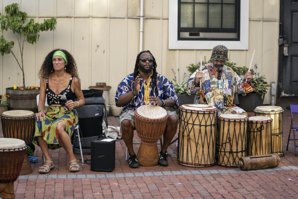 Three seated people playing a variety of African drums, including the djembe and dunun.