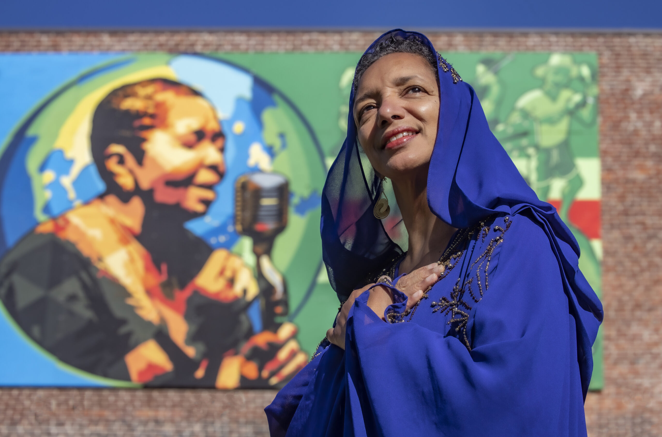 A woman in blue standing in front of a mural of a woman singing.