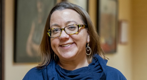 A smiling person of medium light skin tone with short light brown hair, wearing tortoiseshell rimmed glasses and circular earrings, and a dark blue top