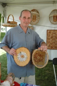 A smiling Native man in a blue button up shirt holding his birchbark art pieces up to the camera