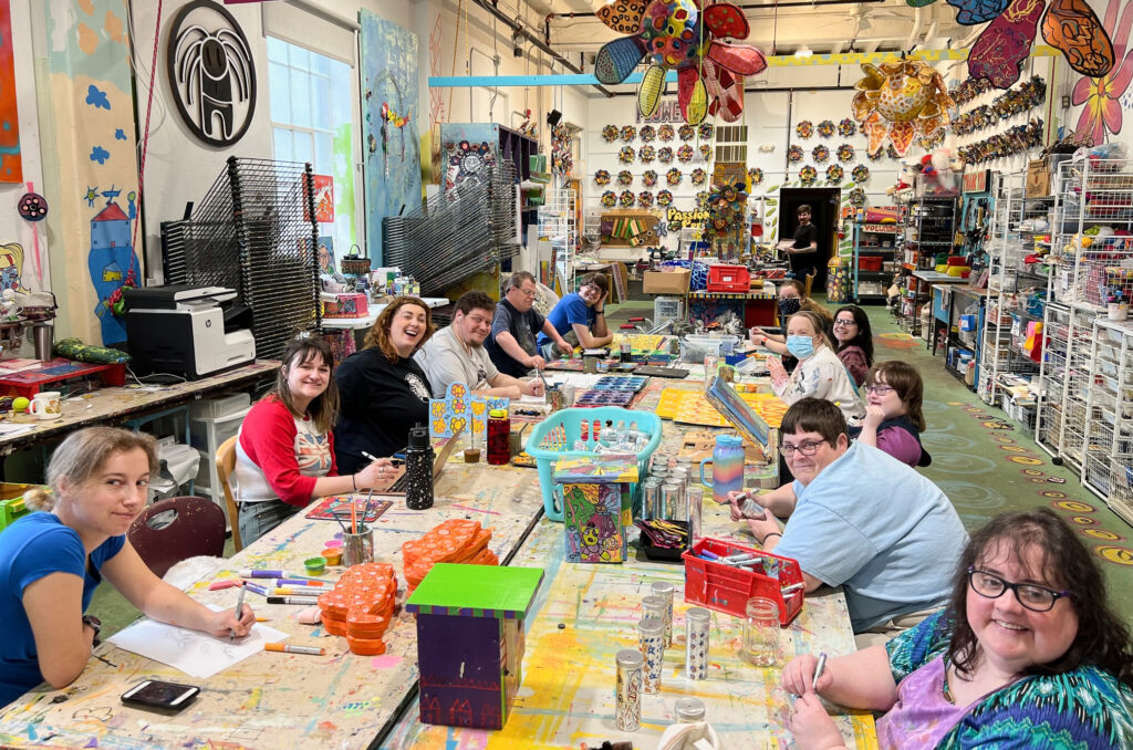 A dozen artists sit around large tables in a big room filled with all kinds of art supplies and artworks. The artists are captured in the middle of working, some with paintbrushes and other instruments in their hands.