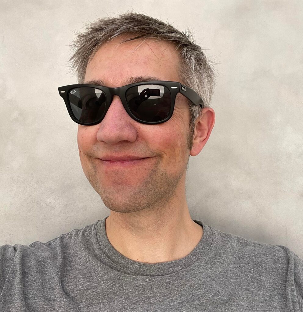 A softly smiling person of light skin tone with short black and grey hair, wearing black sunglasses