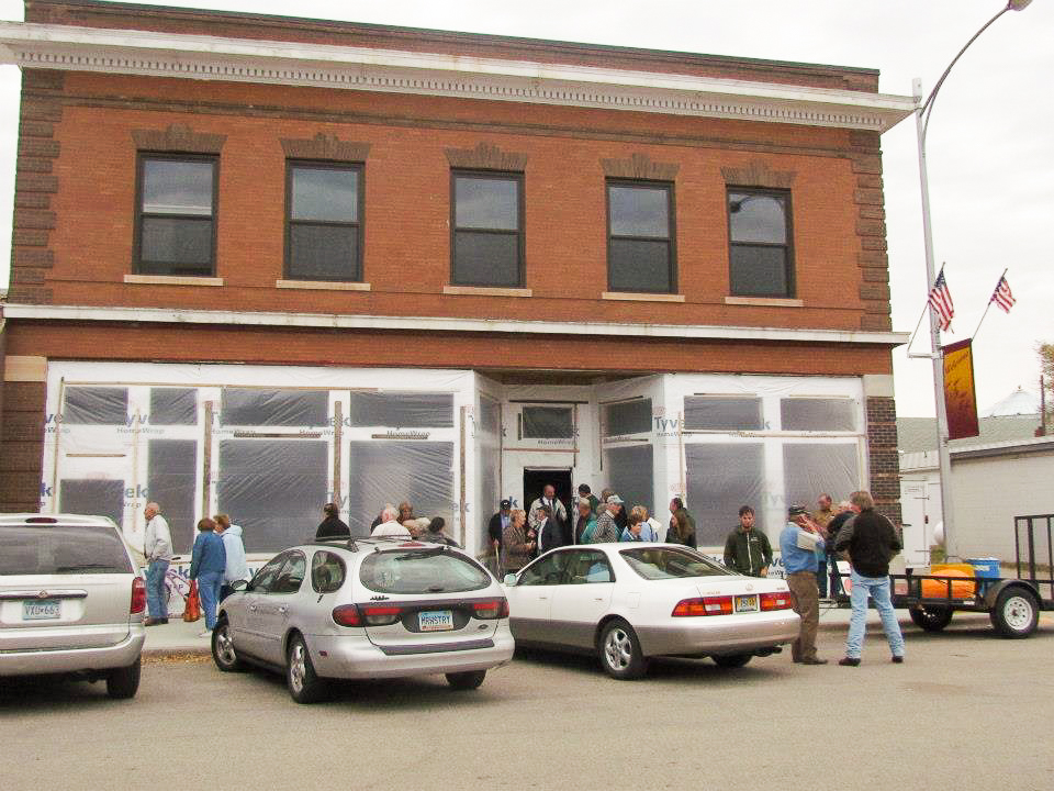 A two-story historic brick building in Maddock, North Dakota, with three parked cars and a dozen people in front of it. The building's first floor with large windows is covered with translucent plastic during renovation in 2013.
