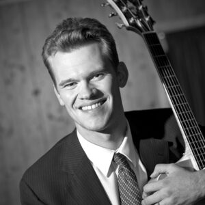A smiling person of light skin tone and short light brown hair, holding a guitar, and wearing a suit and tie combo