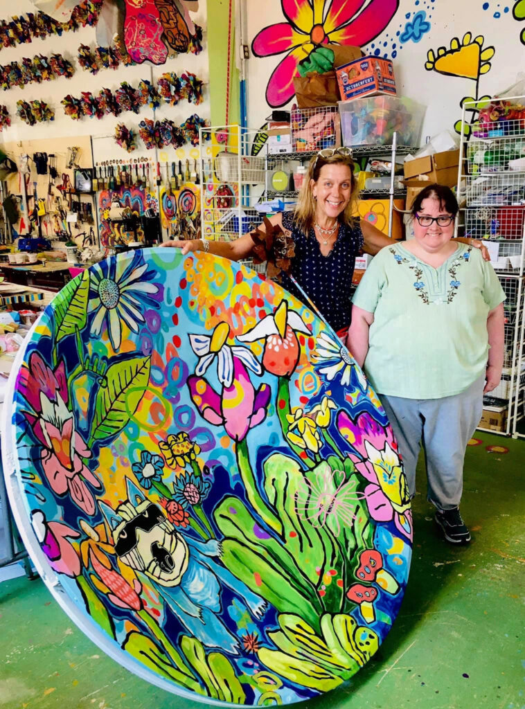 Two people, standing and smiling at the camera, as one holds up a large vintage satellite dish . This satellite dish has been painted by the artist in the photo and it depicts a colorful nature scene with a raccoon hiding behind a bush.