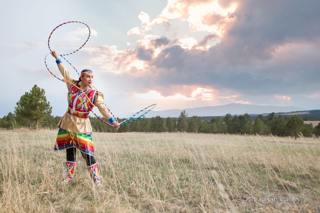 A person of medium light skin tone wearing Native American regalia as a Lakota hoop dancer, holding their hoops poses outdoors. The horizon behind her is a line of trees and a slightly cloudy sunset.